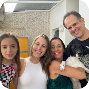 Quatro pessoas de uma família sorriem para foto, onde a mulher segura um cachorro preto e branco no colo