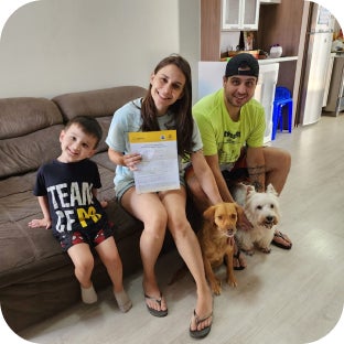 Mulher sentada expondo um termo de adoção em uma das mãos. Em um de seus lados está um homem e um menino do outro lado, todos sorrindo com dois cachorros sentados entre eles.