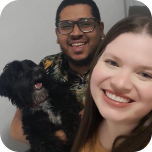 Casal sorrindo para a foto. O homem segura um cachorro preto de pelos compridos que o observa enquanto a foto é tirada