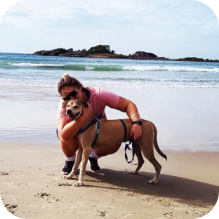 Mulher abraça e beija um cachorro caramelo na areia da praia, com a vista do mar ao fundo