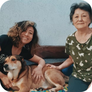 Cachorro caramelo, preto e branco, sentado entre duas mulheres que olham sorrindo para a foto