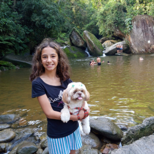 Menina segurando uma cachorrinha peluda caramelo e branco com um lago e pedras ao fundo.