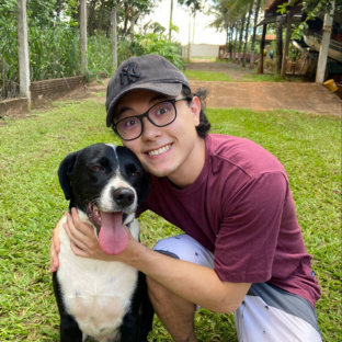 Homem de óculos, boné e camisa vinho, abraçado a um cachorro preto e branco. No fundo, gramado e árvores.