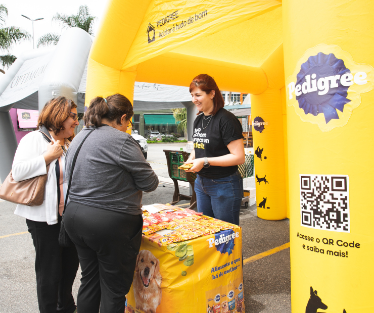 Tenda inflável amarela com logo da PEDIGREE, com uma moça dentro atendendo duas mulheres que escutam mais sobre o programa Adotar é tudo de bom.