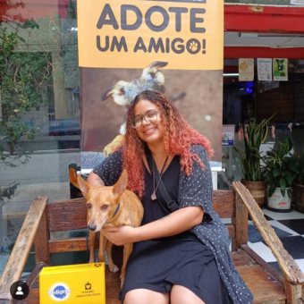 Fotografia da Bárbara sentada em um banco de madeira ao lado do seu cachorro Bob com uma placa 'adota um amigo' ao fundo'.