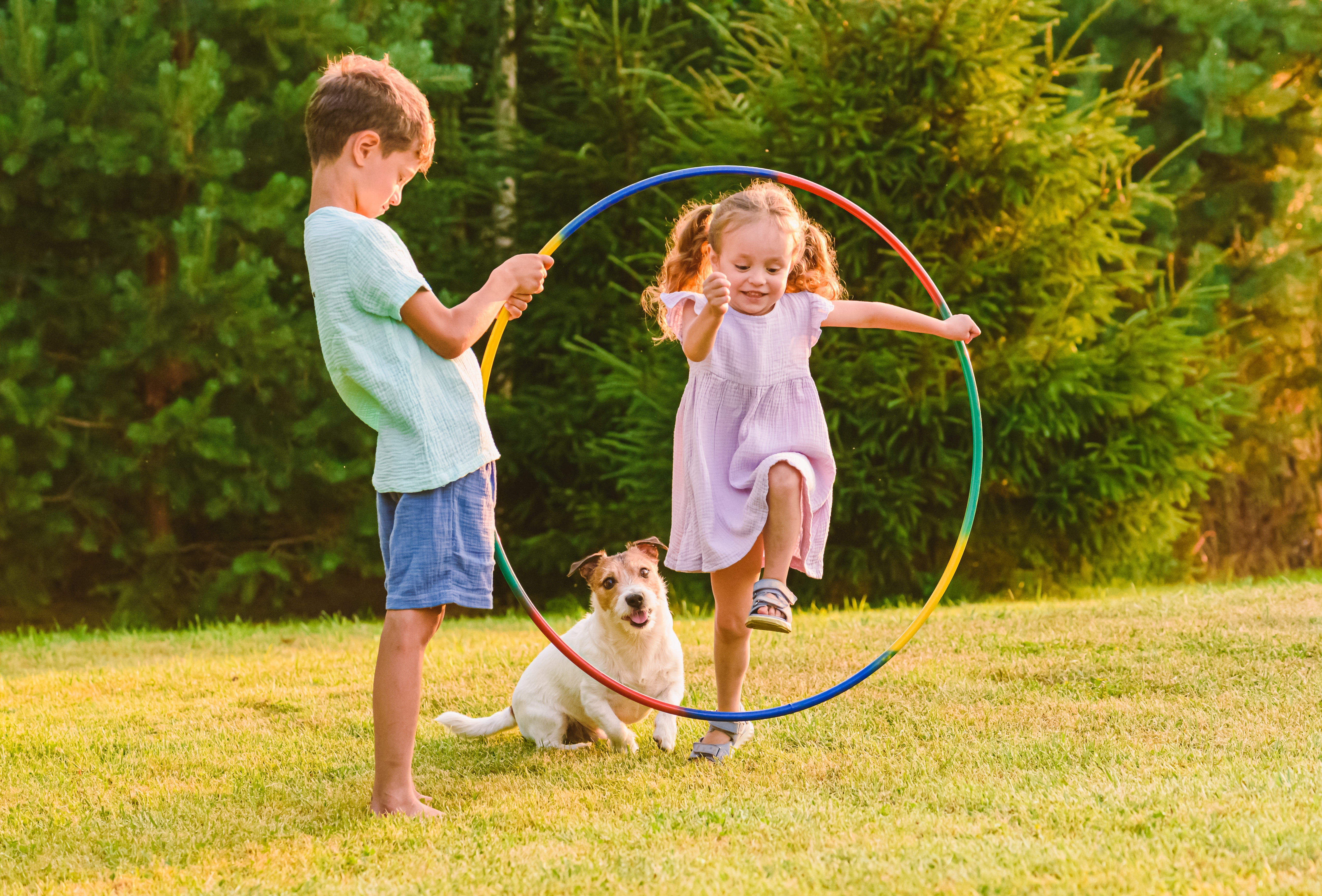 Crianças brincando ao ar livre com um cachorro da raça Jack russell terrier, usando um bambolê colorido para atividades divertidas no gramado.