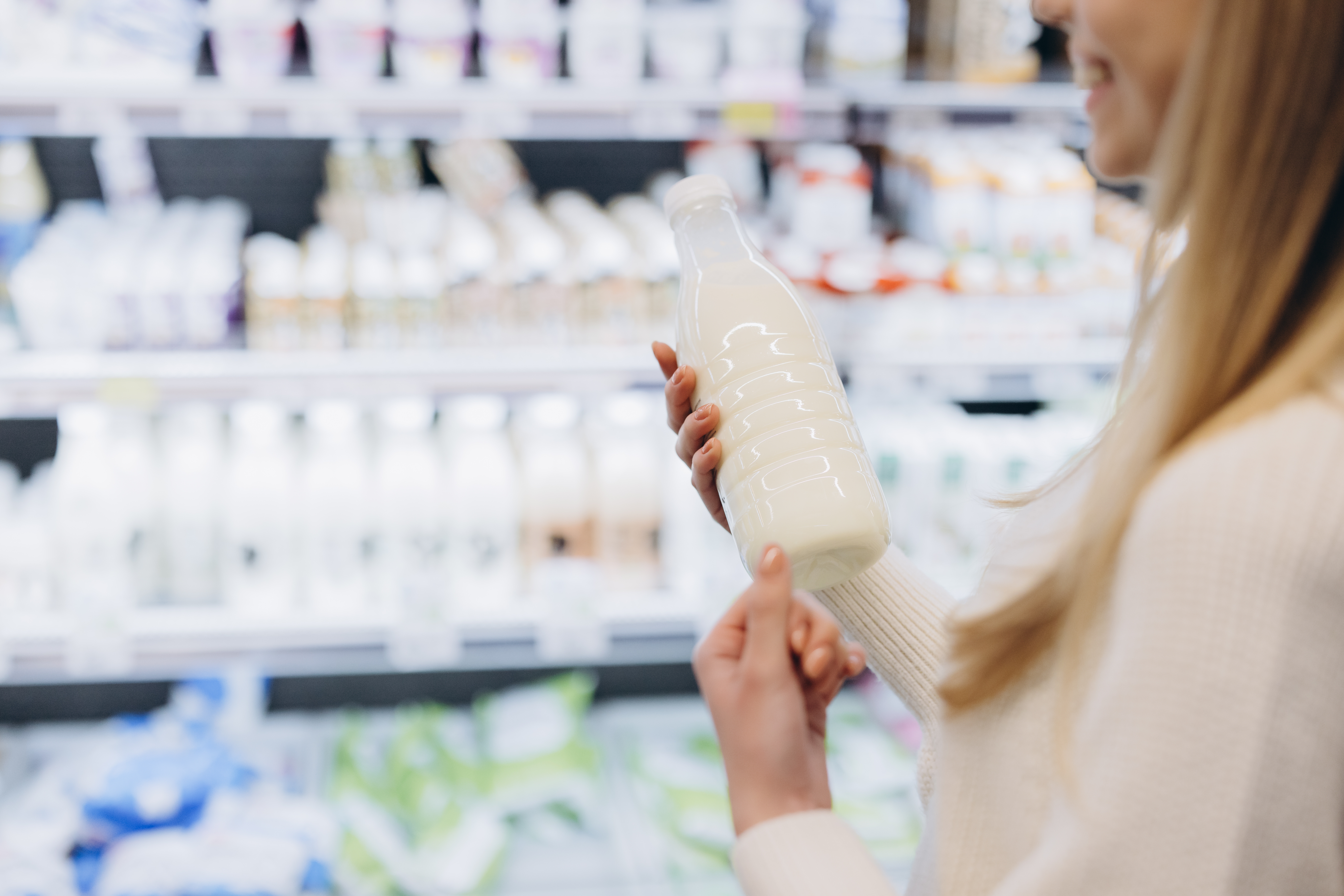 Pessoa segurando uma garrafa de leite em um supermercado, com prateleiras de produtos lácteos ao fundo.