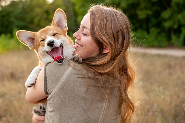 uma mulher segura um cão sorridente no colo, de costas para a câmera. Ao fundo há uma grama alta e escura.