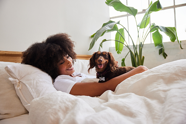 Uma mulher sorri, deitada em uma cama branca, com um cachorro marrom no colo - que também sorri para câmera.