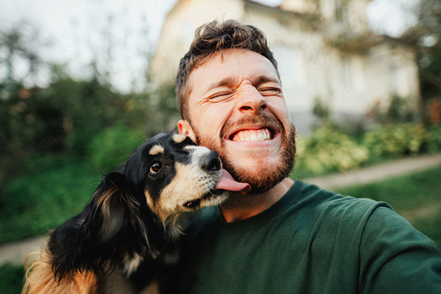 Um cachorro lambe o rosto de um homem, enquanto esse sorri. O fundo está desfocado.